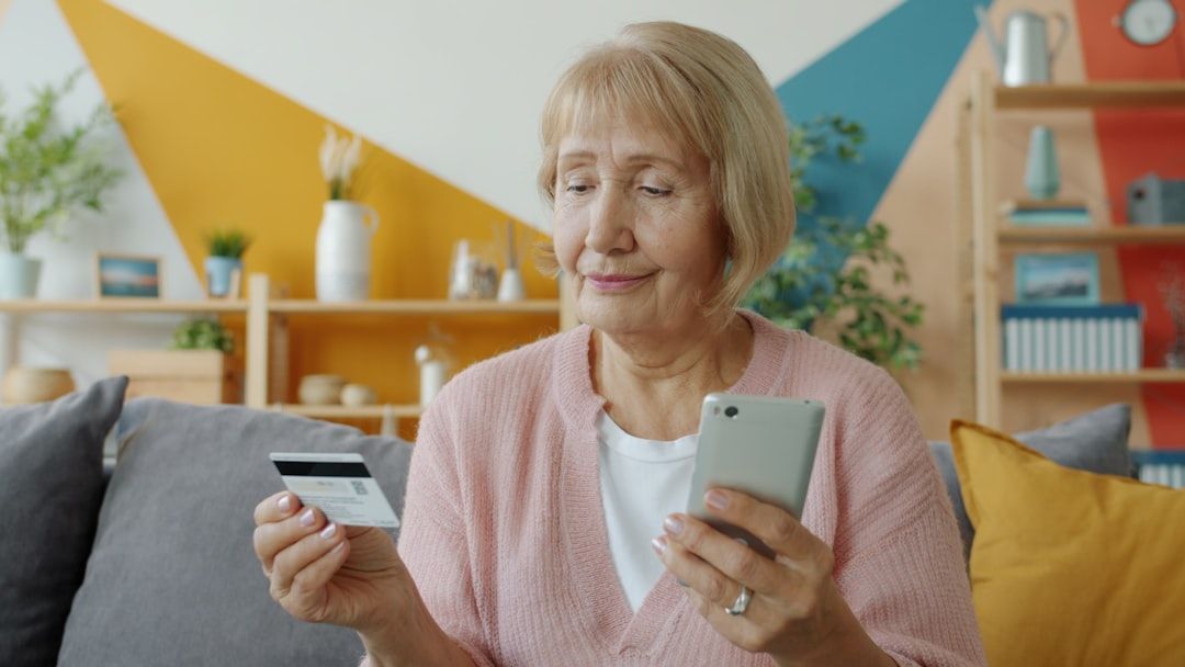 En senior kvinna som använder sin mobiltelefon och ett bankkort i ett vardagsrum.